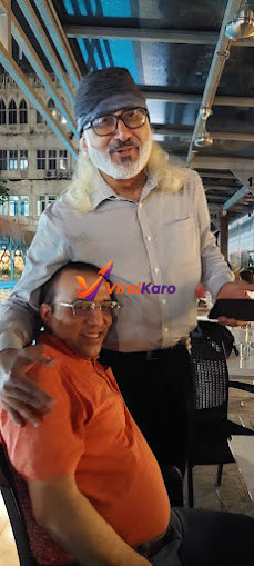 Two men smiling and embracing at a dimly lit outdoor restaurant in Mumbai. The standing man, with a long white beard, glasses, and a beanie, has his arm around the seated man in an orange polo shirt. A historical building with intricate architecture is visible in the background, suggesting a lively urban setting at night.