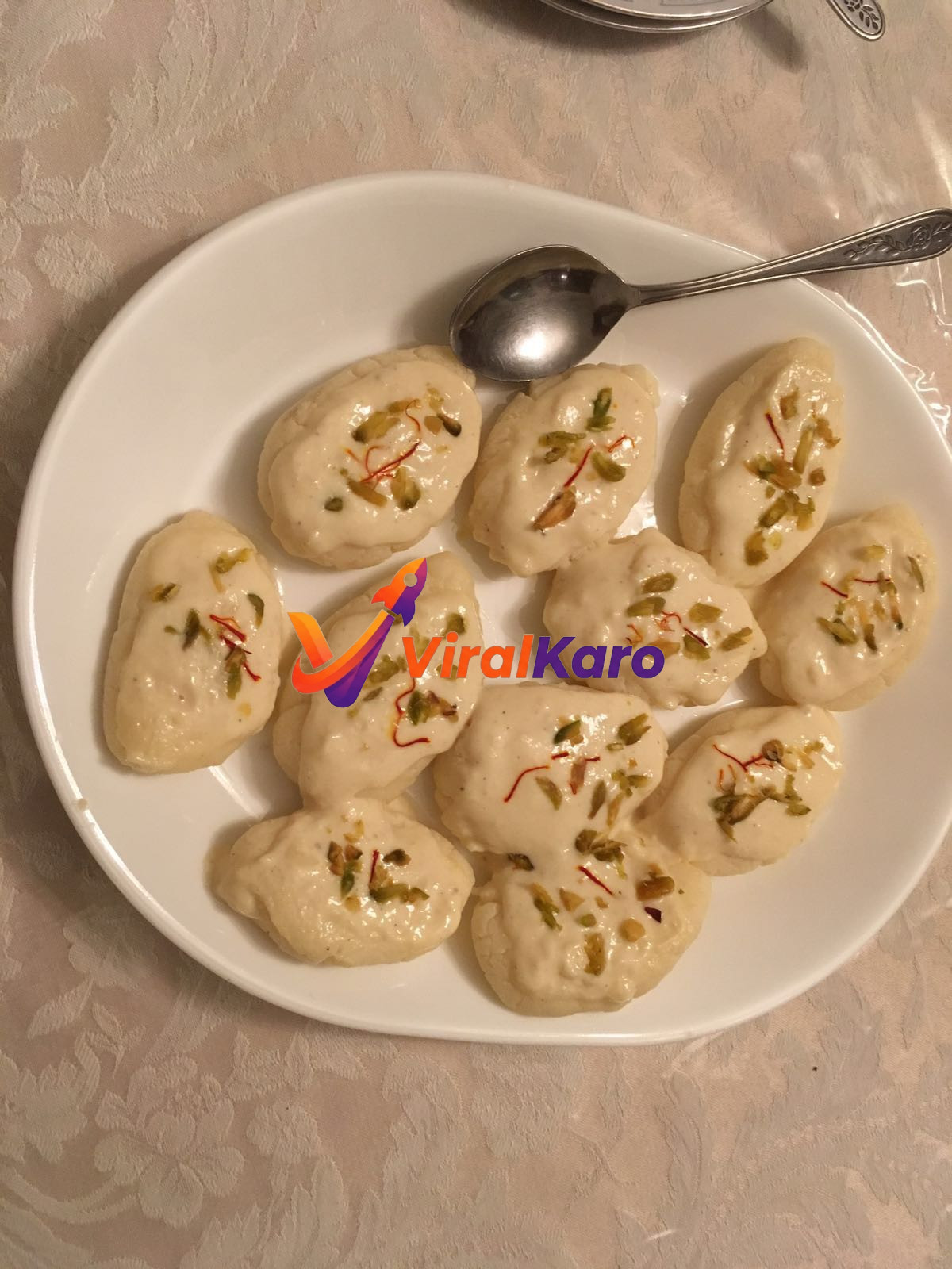 A white plate filled with several pieces of creamy Malai Cham Cham dessert, garnished with saffron strands and chopped green pistachios, with a silver spoon on the side.