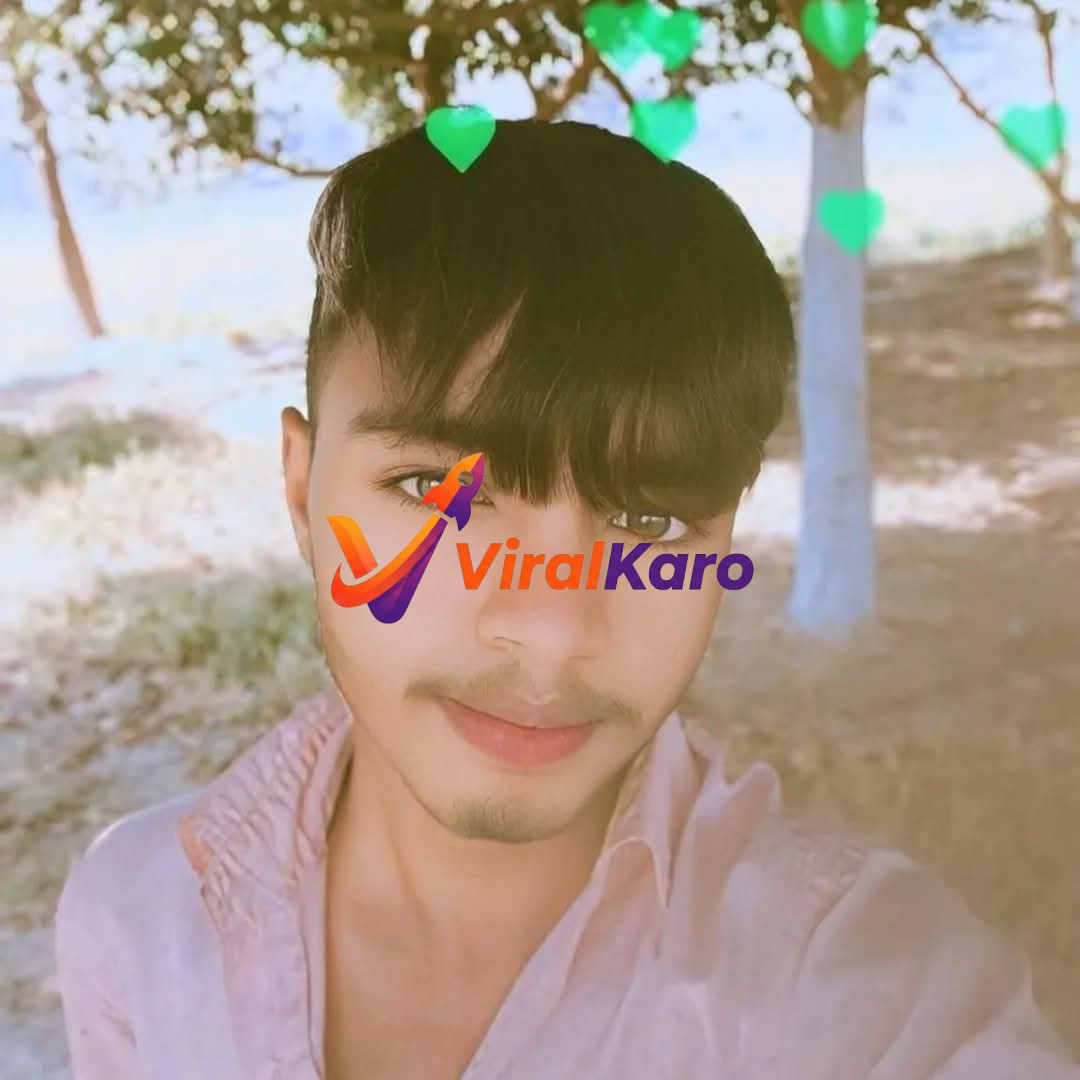 A close-up selfie of a young Indian man with short, dark hair and light brown eyes, smiling subtly. He is wearing a light pink collared shirt and stands outdoors with green heart-shaped digital filters floating above his head. The background shows blurred trees and a bright, natural setting.